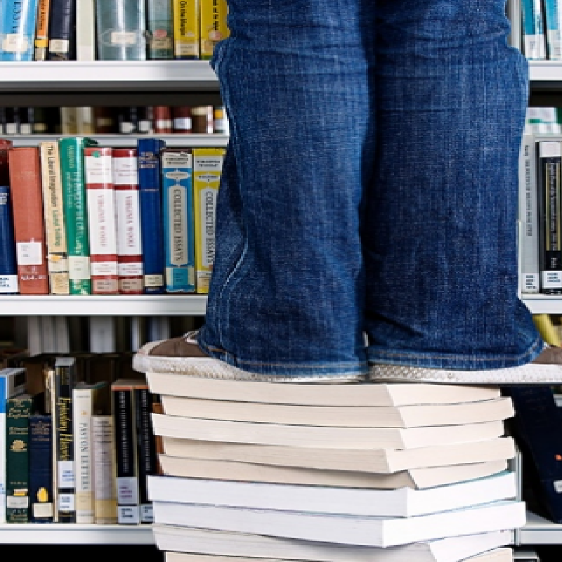 Person in jeans standing on a stack of books in front of a library bookshelf.